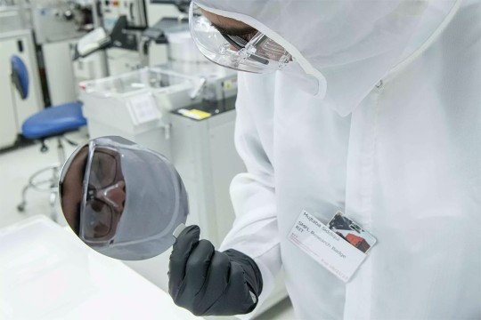person wearing a clean suit holding a semiconductor with tweezers that reflects the image of his face.