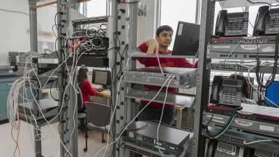 a student working in a server room, with several racks holding communications equipment.