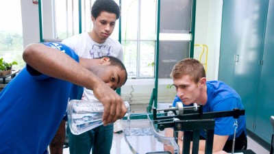 Students pouring a beaker of water into a larger glass container.