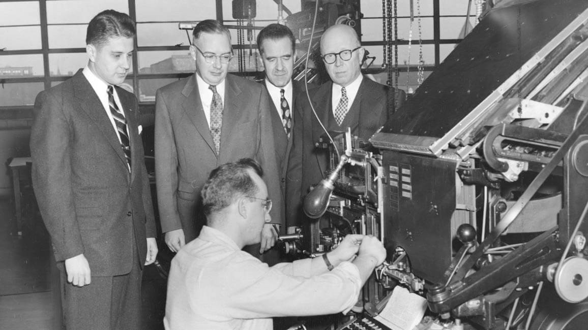 Men in suits standing behind a person using a large piece of equipment.