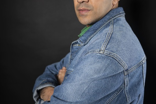 Studio portrait of a man in clear brown glasses and a blue denim jacket, arms crossed as he looks over his shoulder with a calm, confident expression.