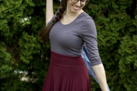 Smiling woman with glasses and a braid twirls outdoors, wearing a gray top and burgundy skirt, her blue scarf flowing against green hedges.
