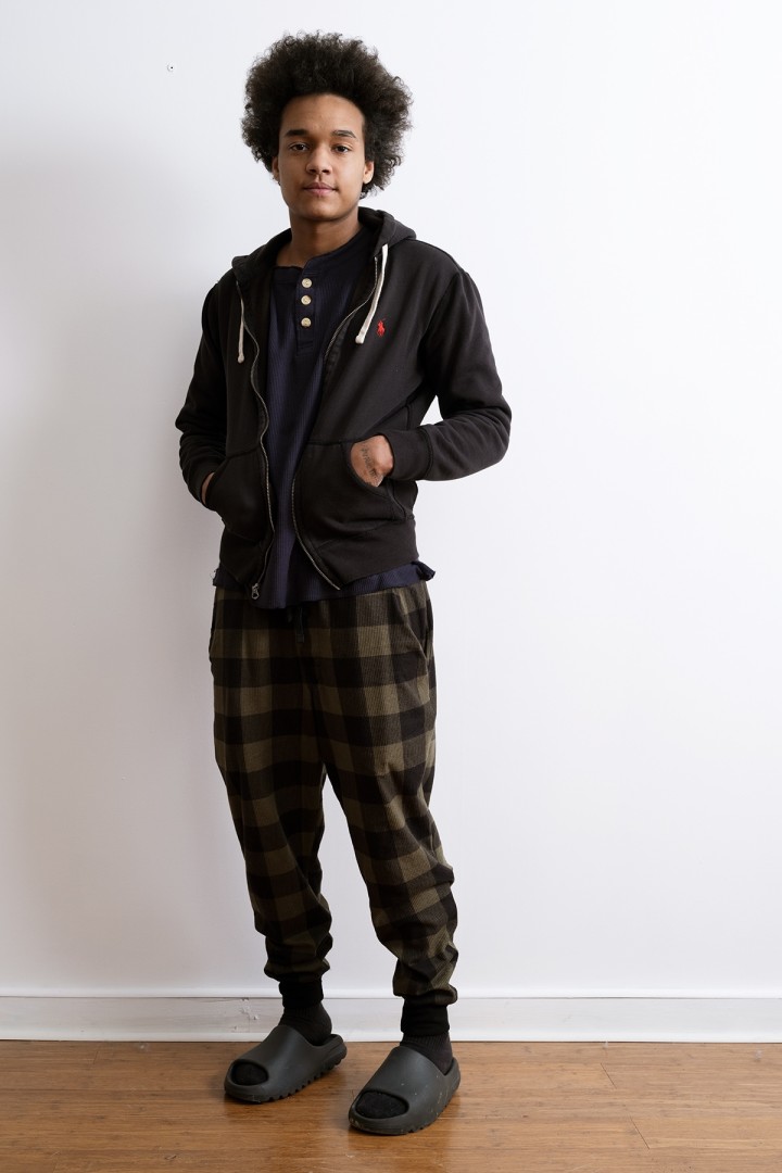 an image of a person wearing dark clothing, hands in pockets, standing against a white wall.