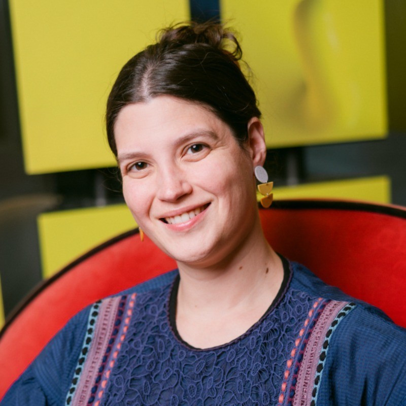 “Smiling woman with dark hair in a loose bun wearing a patterned navy top and geometric earrings, seated in a red chair with a bright yellow and black abstract background.”