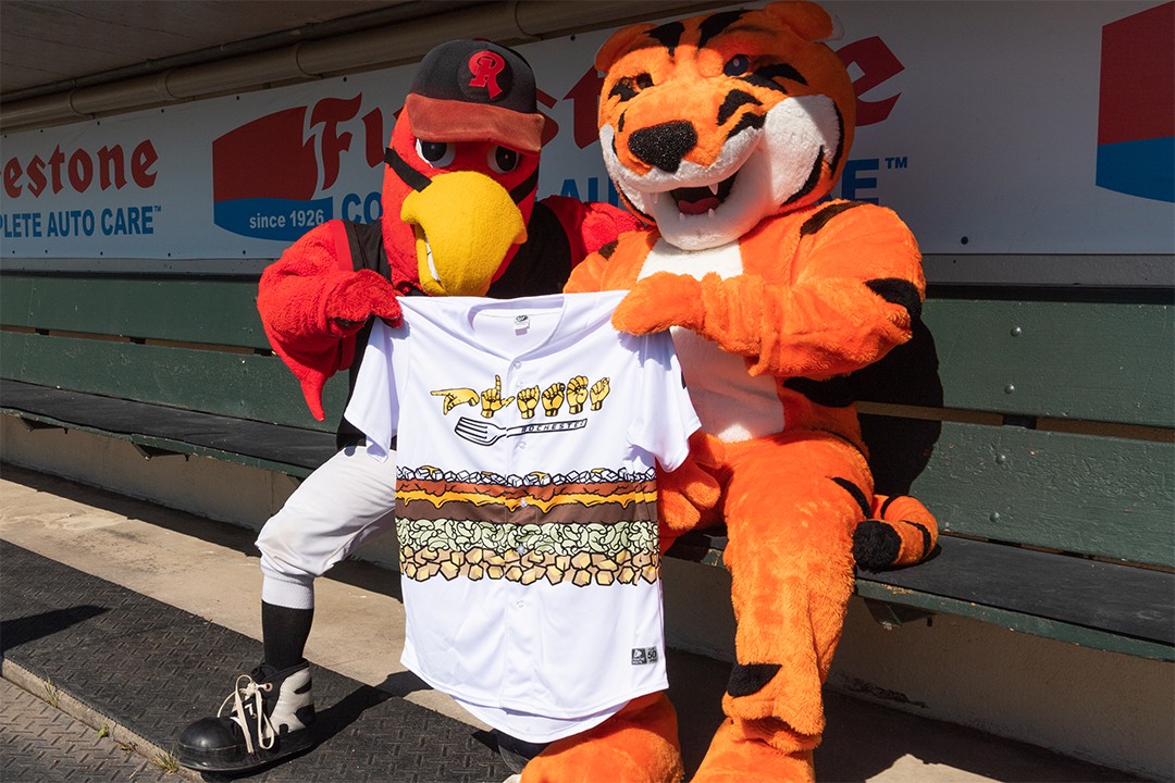 Mascots Spikes and RITchie are sitting in a baseball dugout holding a baseball jersey that reads "Plates" in sign language.