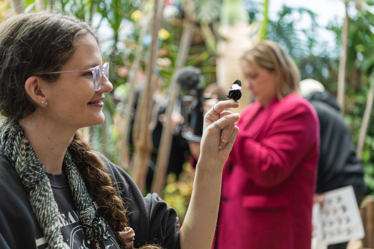Image of a female presenting person wearing glasses and a scarf is holding out their index finger where a butterfly has landed. In the background, other people are milling around indoors among tropical plants. 