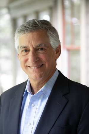 A professional headshot of an older man with short gray hair smiling at the camera. He is wearing a navy blazer over a light blue button-down shirt. The background is softly blurred, showing a bright outdoor walkway with windows and white columns.
