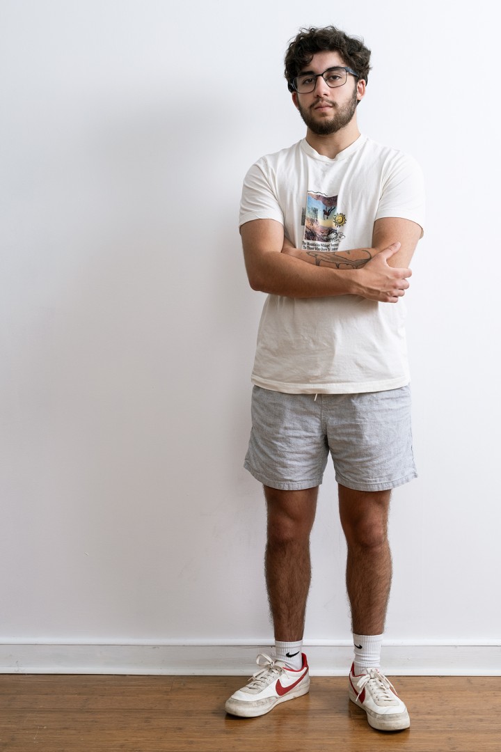 image of a person wearing a white t-shirt and grey shorts, with crossed arms and standing against a white wall.