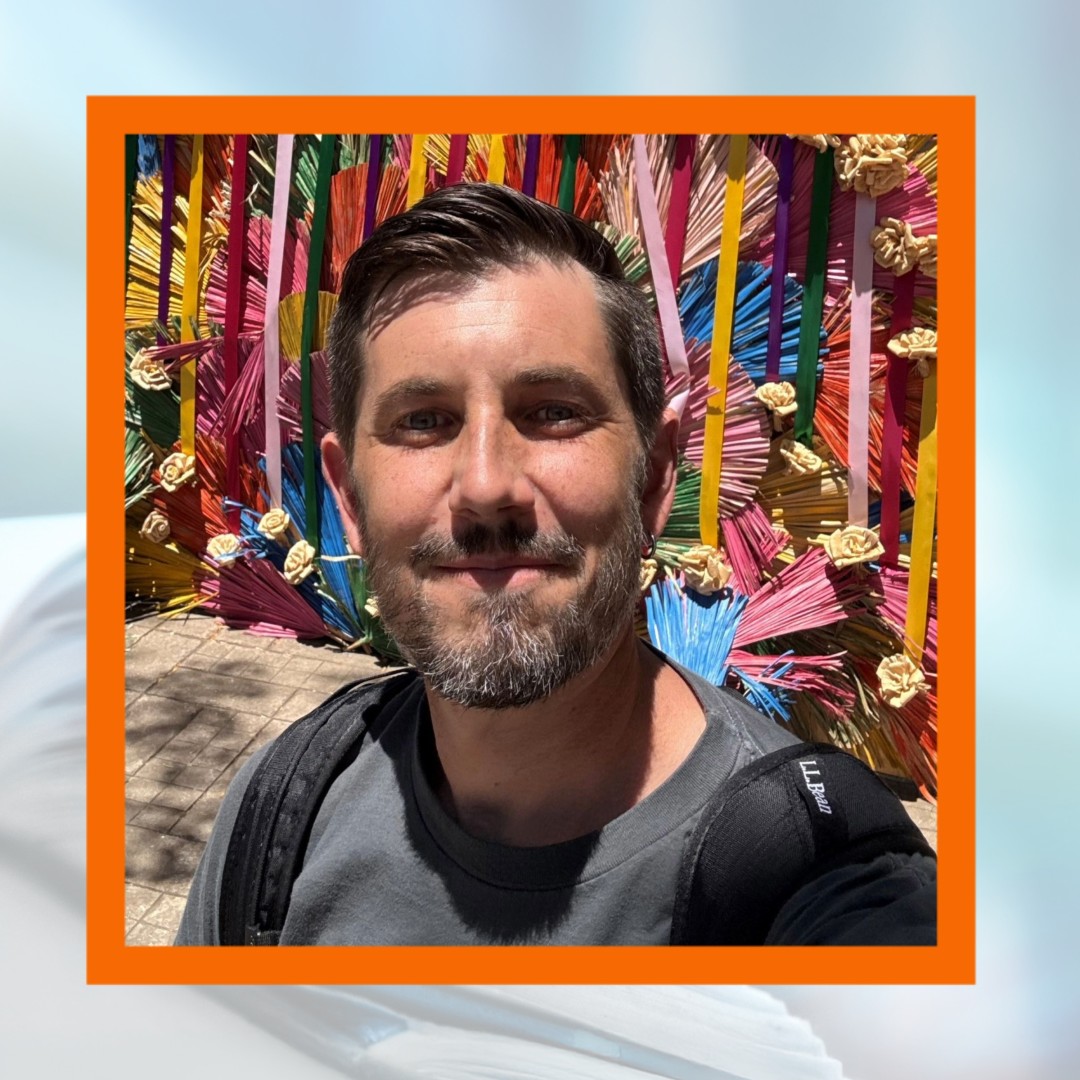 A man with short brown hair, a trimmed beard, and a backpack takes a selfie outdoors in front of a vibrant, colorful art installation made of fan-shaped pieces in red, blue, yellow, pink, green, and orange, decorated with cream-colored flowers. Trees and lamp posts are visible in the background under a sunny sky.