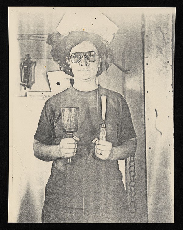 Black-and-white photograph of Dr. Ellen Swartz standing indoors, holding a tool in her right hand and a tool in her left. She wears glasses and a short-sleeved shirt, looking directly at the camera. The background includes workshop elements such as tools, a lamp, and a chain hanging on the wall.