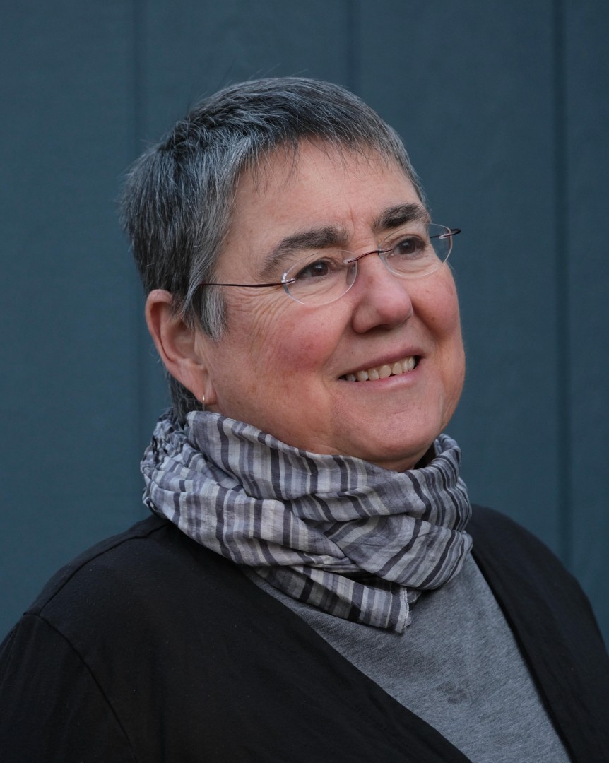 Blue background with a lady with short grey hair and glasses wearing a black sweater and multi grey colored scarf.