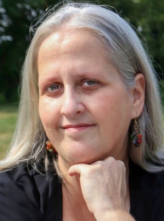 Close-up portrait of a person with long, straight gray hair, resting their chin on their hand. They are outdoors with a softly blurred green background, wearing a dark top and small earrings, and looking calmly toward the camera.