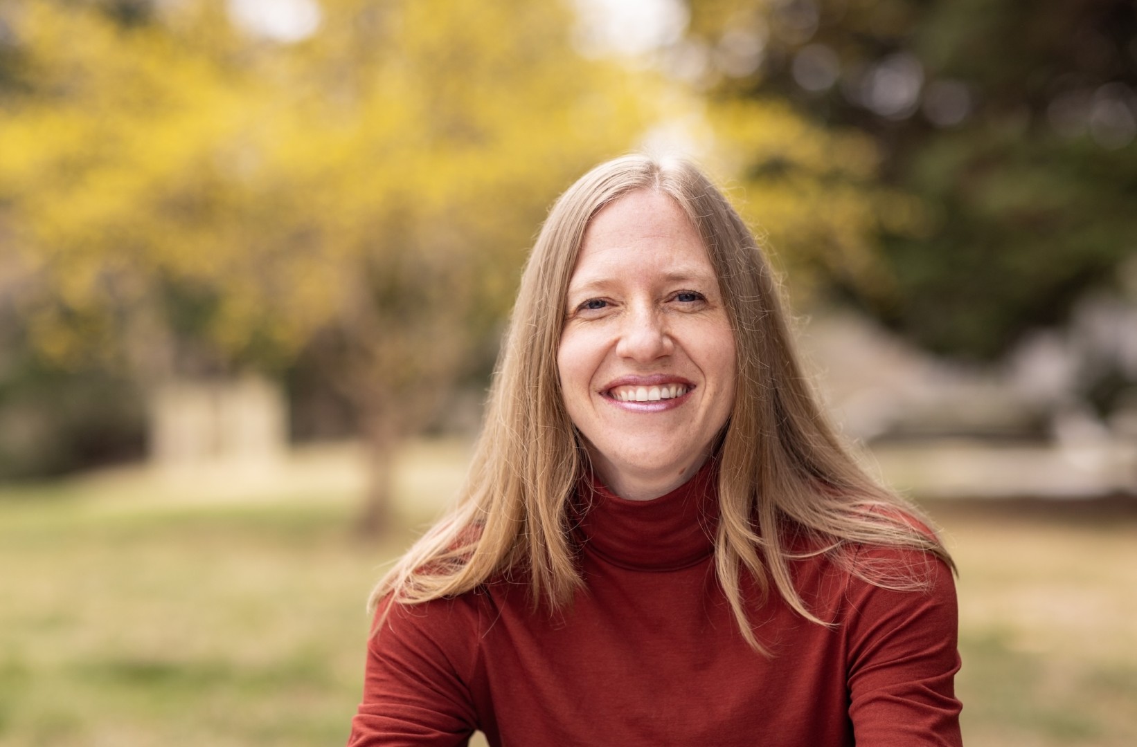 Picture of young woman with blonde hair, wearing a red turtleneck with blurry background.