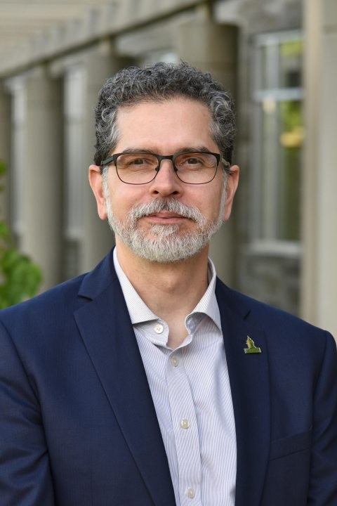 A professional headshot of a man with short, curly salt-and-pepper hair and a neatly trimmed gray beard and mustache. He is wearing rectangular glasses, a navy blazer, and a light-colored button-down shirt, with a small green lapel pin on his jacket. He stands outdoors in front of a modern building with large windows and stone columns, with greenery softly blurred in the background. He faces the camera with a slight smile.