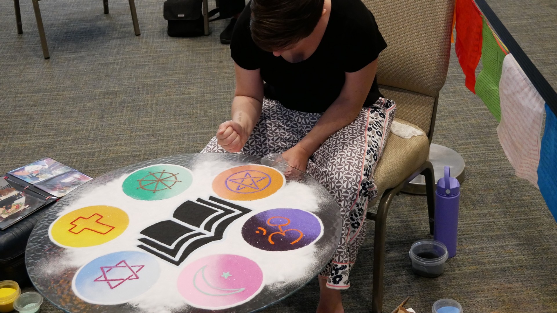 woman sits performing sand art, creating a mandala on a table with various spiritual/religious symbols