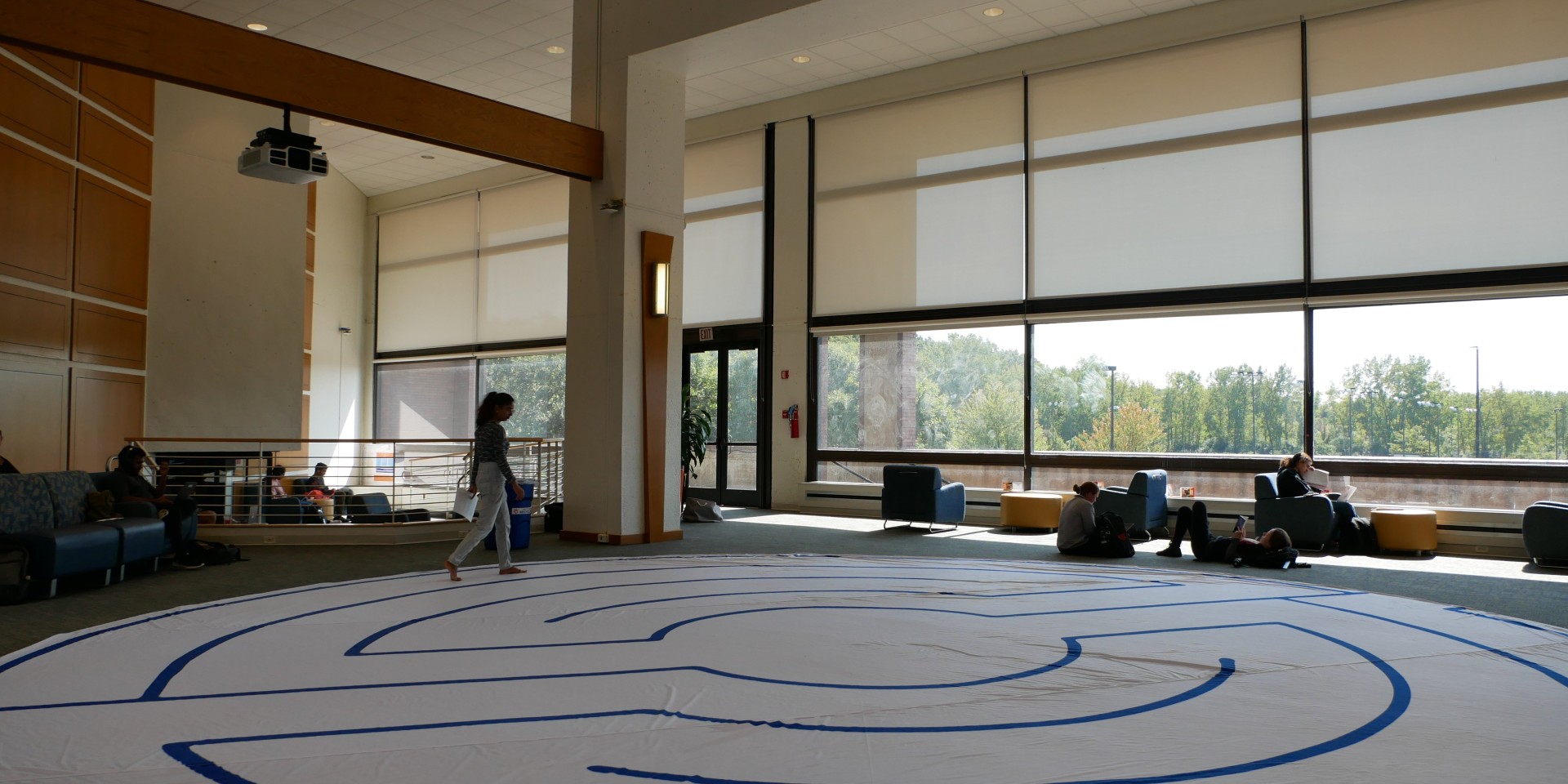 woman walking labyrinth on floor of large open room (with large windows) with students studying in the background