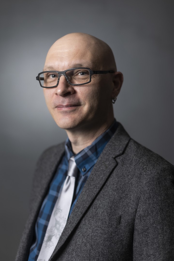 Image of the speaker Matthew Wolf-Meyer in a dark grey suit jacket, blue plaid shirt, silver tie and a grey background
