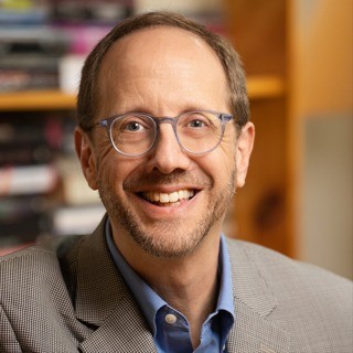 A smiling middle-aged man with short light brown hair and a neatly trimmed beard, wearing round eyeglasses, a blue button-down shirt, and a light gray blazer. He is facing the camera in a warmly lit indoor setting, with softly blurred shelves and books in the background.