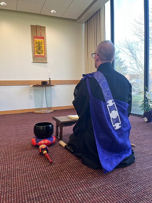 Buddhist priest in robe kneeling and meditating in chapel space with traditional Nichiren Buddhist paraphernalia surrounding him