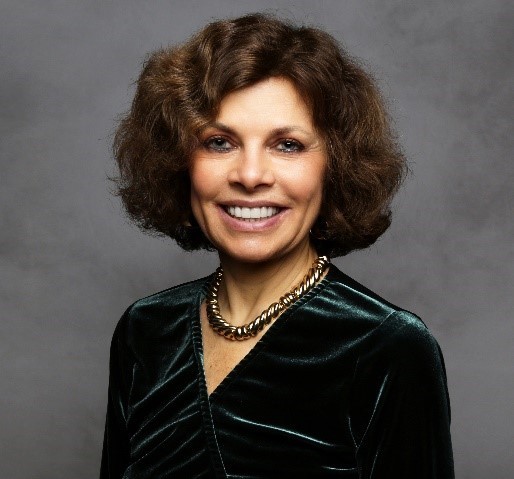 A woman with short curly brown hair is smiling at the camera. She is wearing a dark green velvet top and a gold chain necklace, posed against a gray studio background.