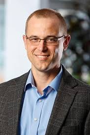 Professional headshot of a smiling man with closely cropped hair and glasses, wearing a blue button-down shirt and a gray blazer. He is facing the camera against a softly blurred indoor background.
