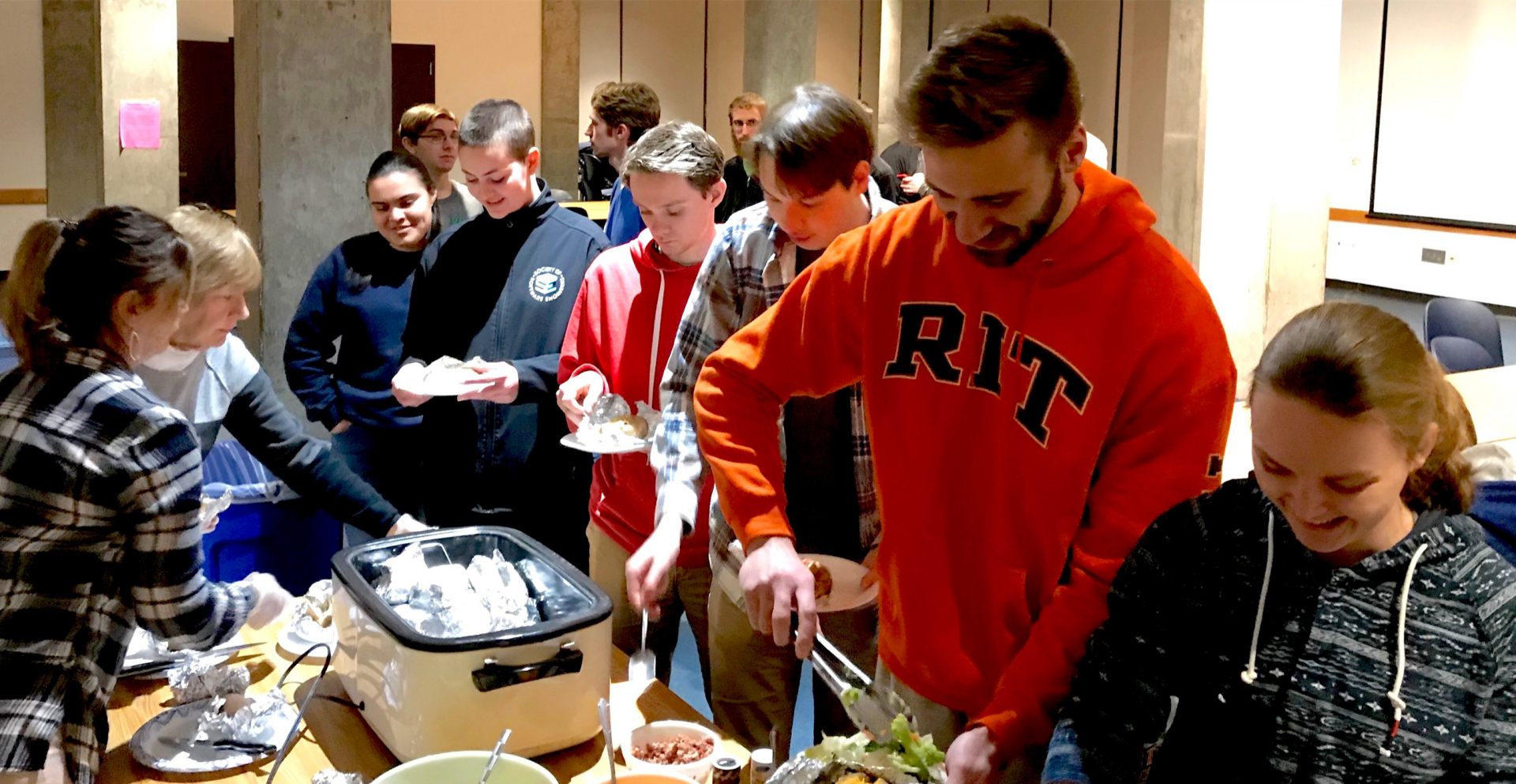 A photo of people standing in line for food. They are wearing a variety of hoodies and sweaters, everyone is smiling.
