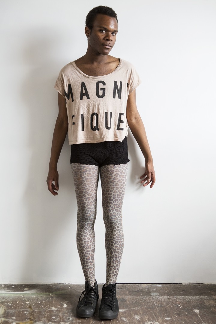 a person wearing a white shirt with bold graphic letters and black shorts standing against a wall.