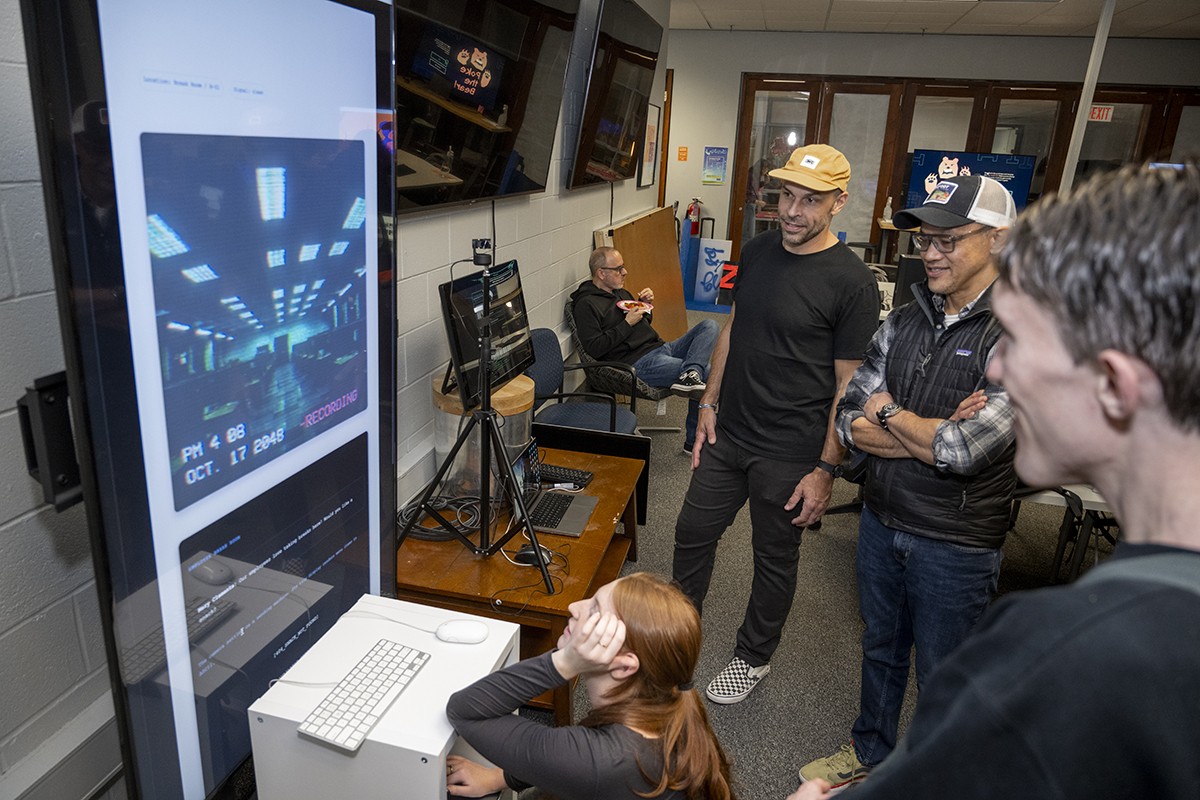 Students and faculty smile as they look at a digital experience on a large screen.