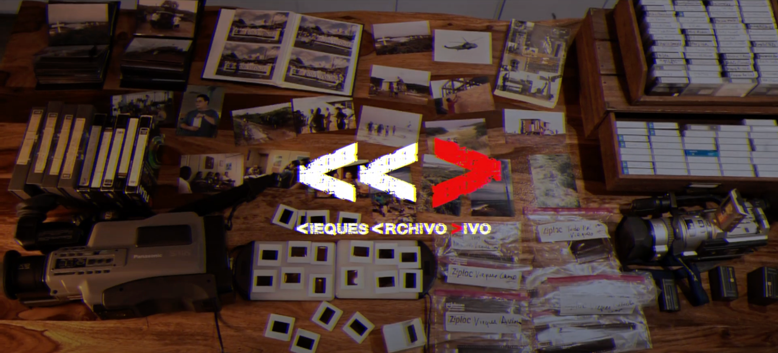 A wooden table covered with old media formats and archival materials, including VHS tapes, photographs, film slides, camcorders, and labeled plastic bags filled with smaller tapes. At the center, a graphic with stylized arrows and glitch effects reads “<< VIEQUES <ARCHIVO> IVO >>” in white and red text.