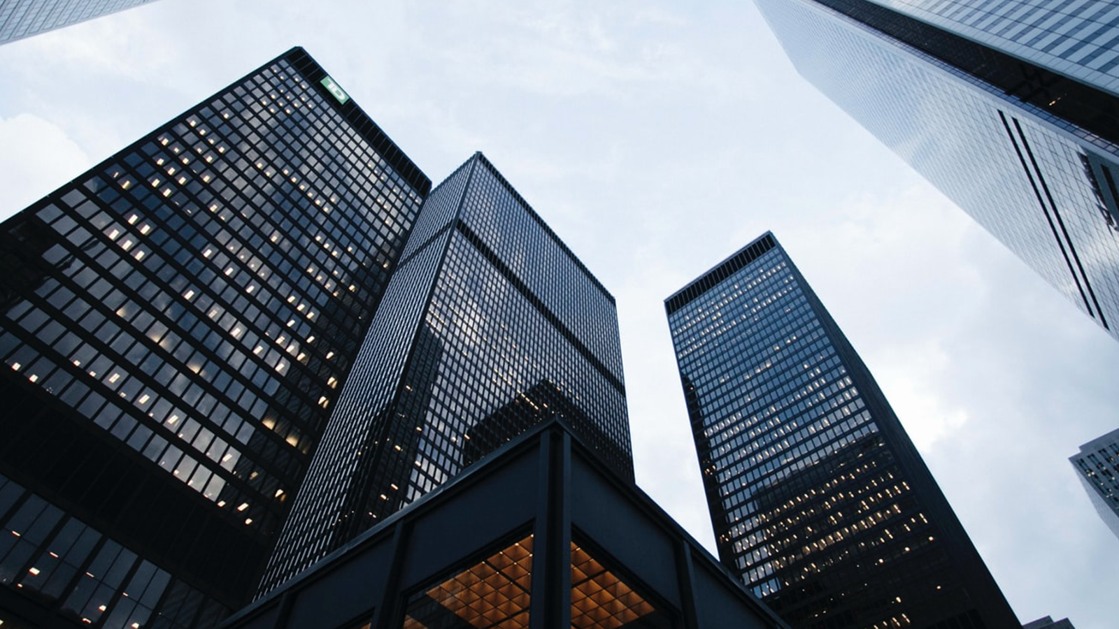 A view of several tall skyscrapers with reflective glass facades reaching into a blue sky with light cloud cover, seen from ground level. One skyscraper has a green logo near the top.