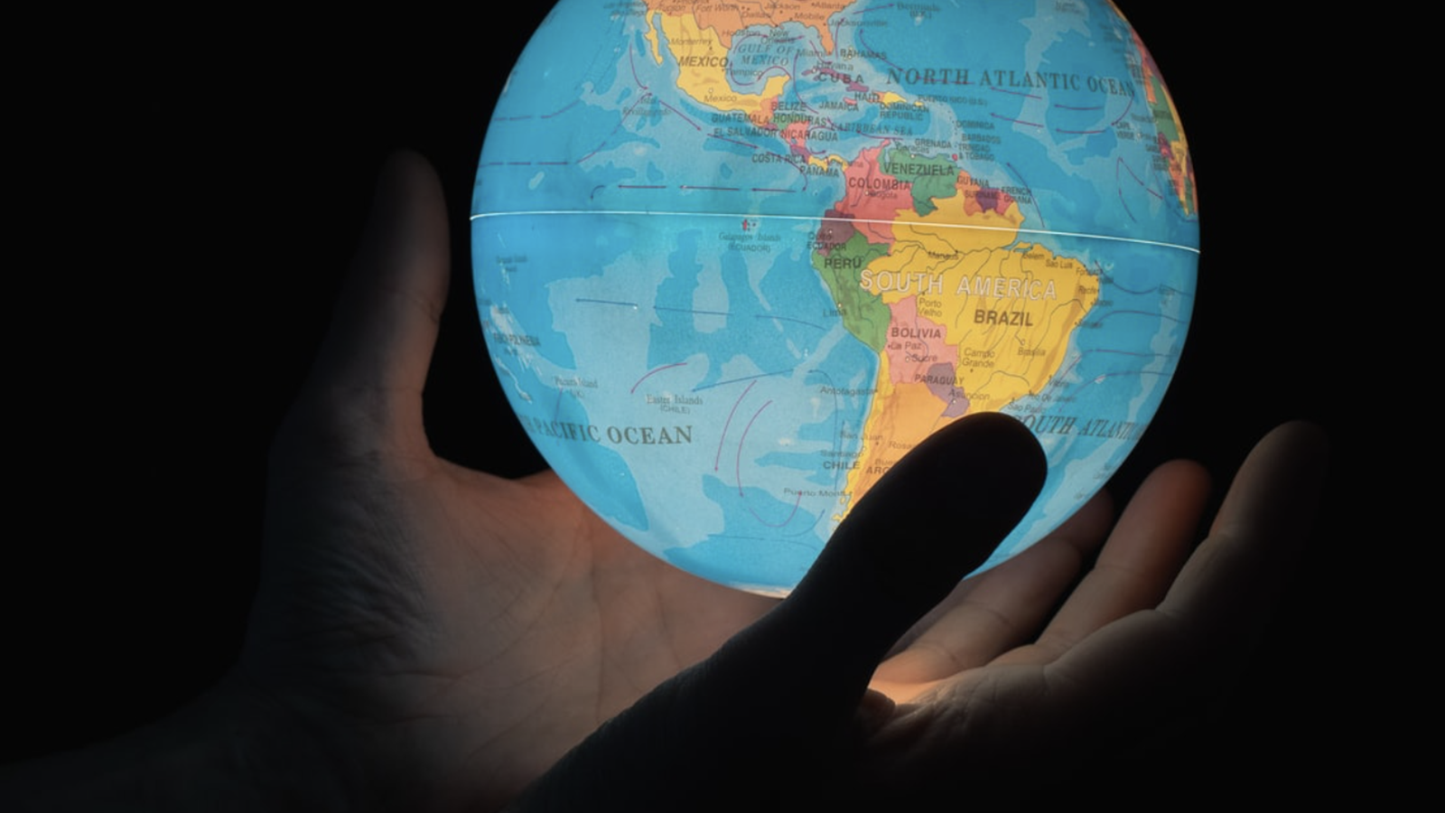 A hand holding a small, illuminated globe against a dark background, highlighting the continents with a focus on Central and South America.
