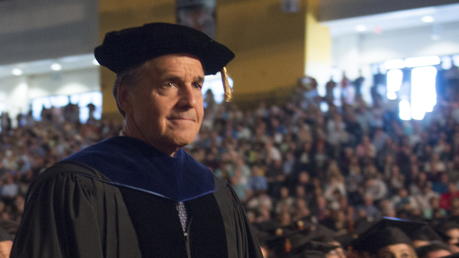 RIT's Senior Vice President wearing a graduation cap and gown, standing in front of a large audience at a commencement ceremony.