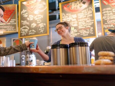 A Java's employee serving a coffee to a customer, with bagels atop the counter