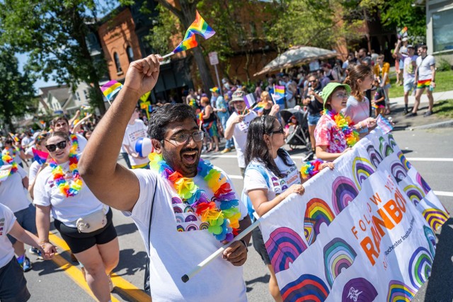 Many people walk on the street at a parade wearing rainbow items and waving rainbow flags.