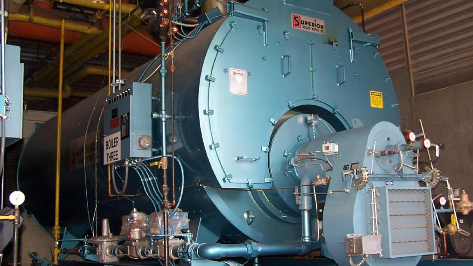A large industrial boiler with various pipes and gauges, labeled “Superior Boiler Works, Inc.” The boiler has a prominent front door with a circular window and control boxes on the left side, situated inside an industrial facility.