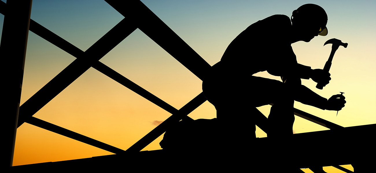 A man, kneeling on the frame of a building, holding a hammer and about to drive a nail.  He is silhouetted by the sunset behind him. 