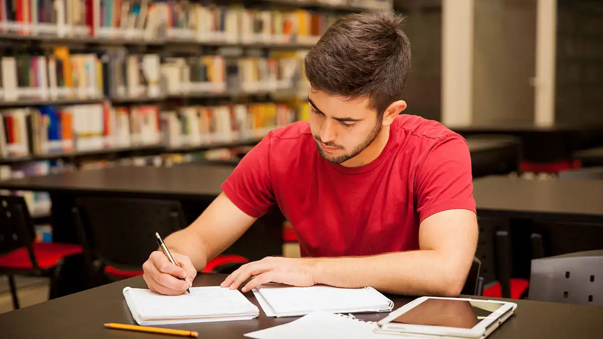 a college student seated at a table with a tablet and notebooks.