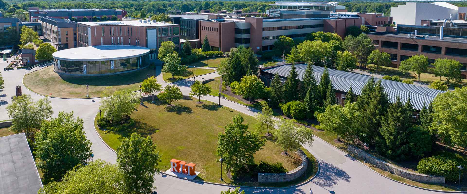 Aerial view of R I T campus, with brick and glass buildings, grassy and treed areas, and an orange R I T statue.
