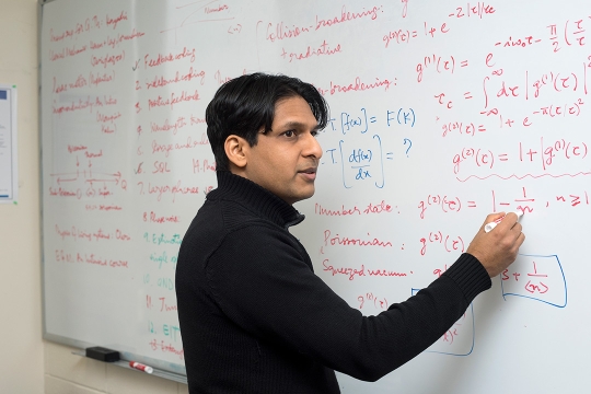 Professor wearing black turtleneck sweater writes equations on dry-erase board.