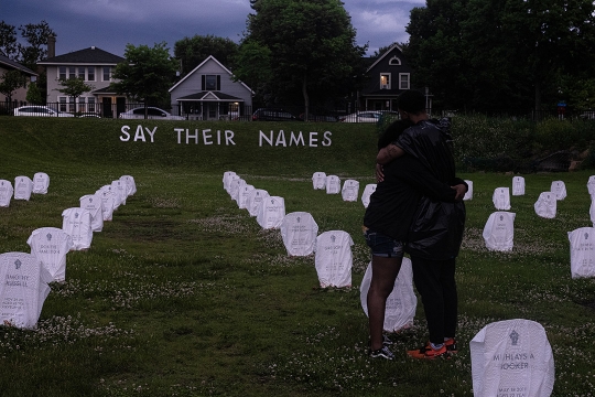 two people hugging in field with makeshift grave markers and a sign that reads "Say their names."