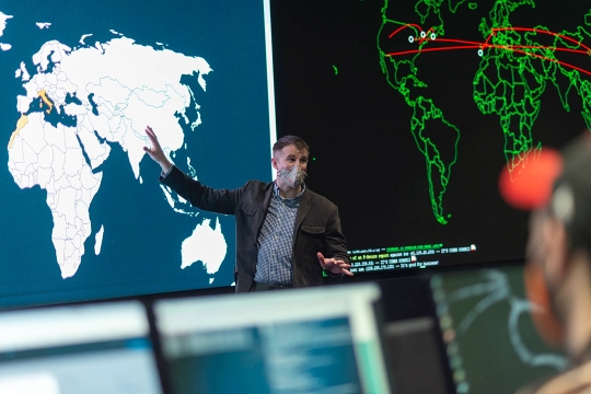 professor wearing a facemask and gesturing toward a world map.