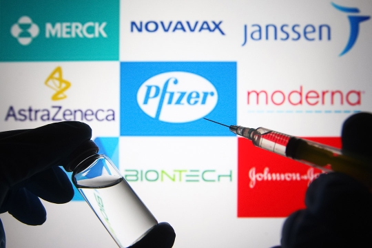 syringe in foreground and logos and names of different vaccine producers in the background.