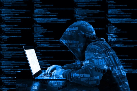 person wearing a dark hoodie typing on a laptop with an overlay of cybersecurity-related words.