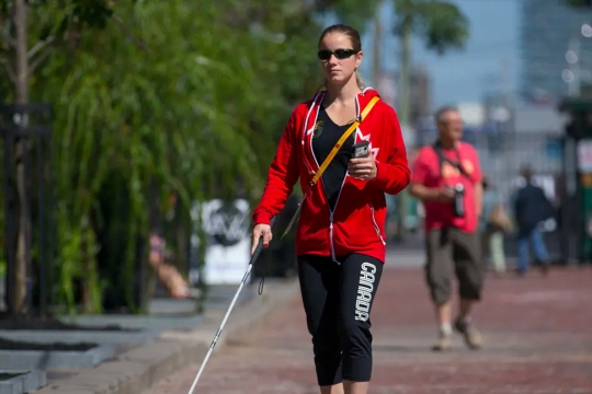 person walking wearing sunglasses, holding a smartphone, and using a guide cane.