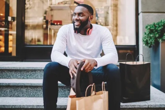 man sitting on steps outside of a store with paper shopping bags.