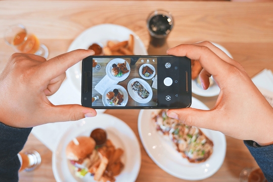 a person taking a photo of four plates of food with a smartphone.