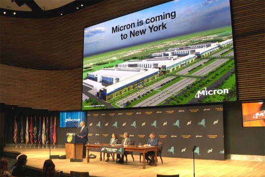 One official standing at a podium and three sitting at a table on a large stage with a large screen behind them that says, "Micron is coming to New York"