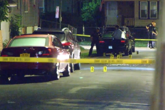 crime scene at night with police tape surrounding part of a street and numbered cards and broken glass on the ground.