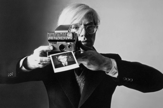 Andy Warhol holding a Polaroid camera that's printing a photo of himself.