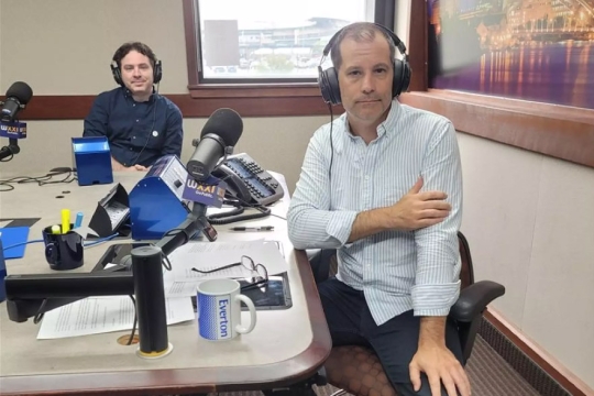 two men wearing headsets and sitting around a table in a radio station.